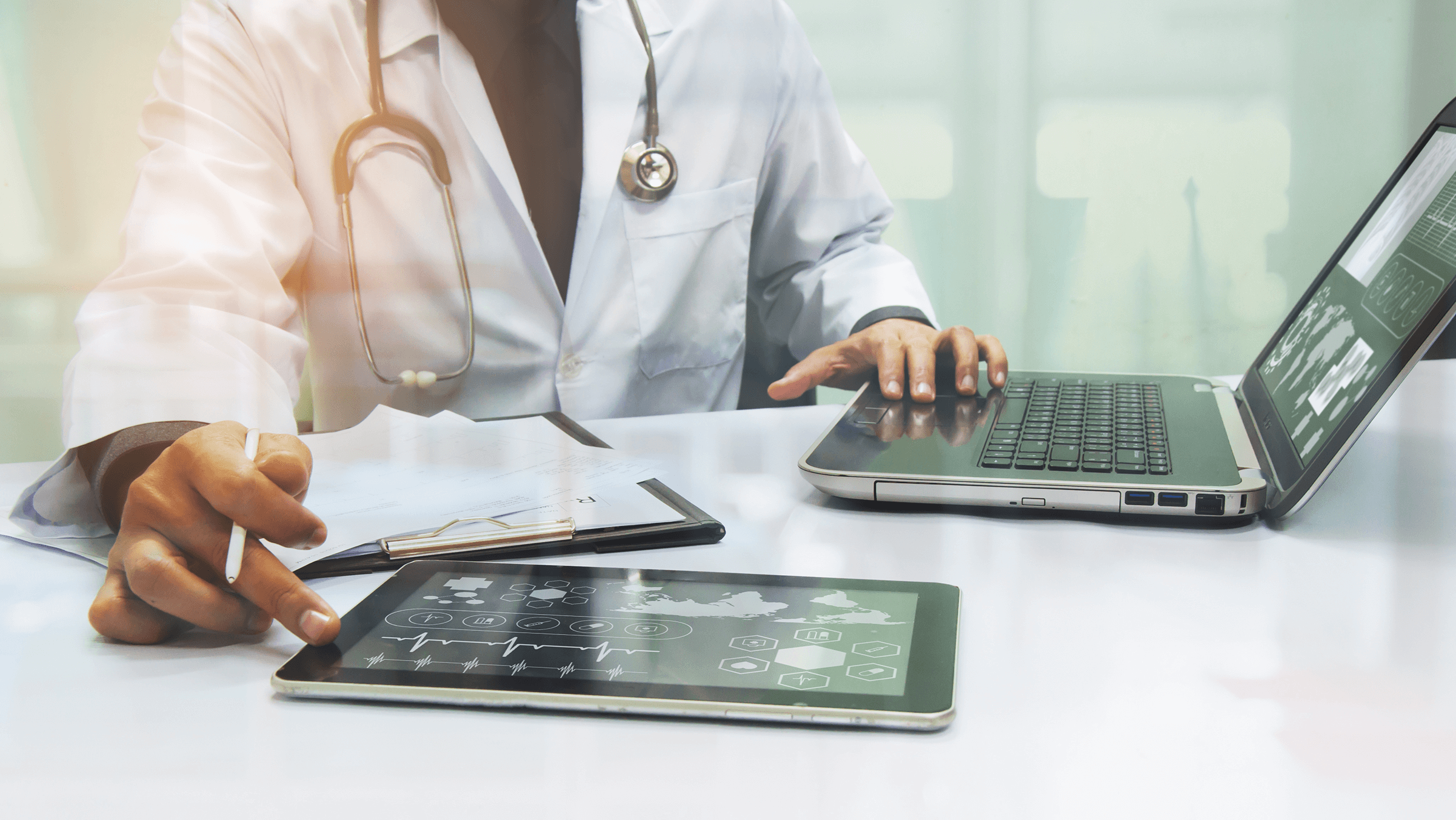 doctor in white coast working on laptop and tablet while taking notes