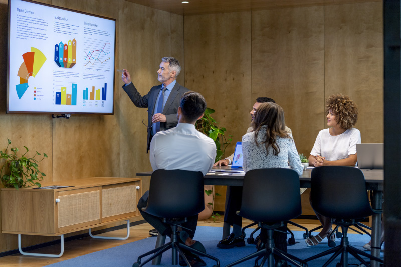 Mature man giving a big data presentation on a tv in a board room. There are several financial graphs and charts on the screen with a diverse group of people in the meeting room. There is paperwork and technology on the table